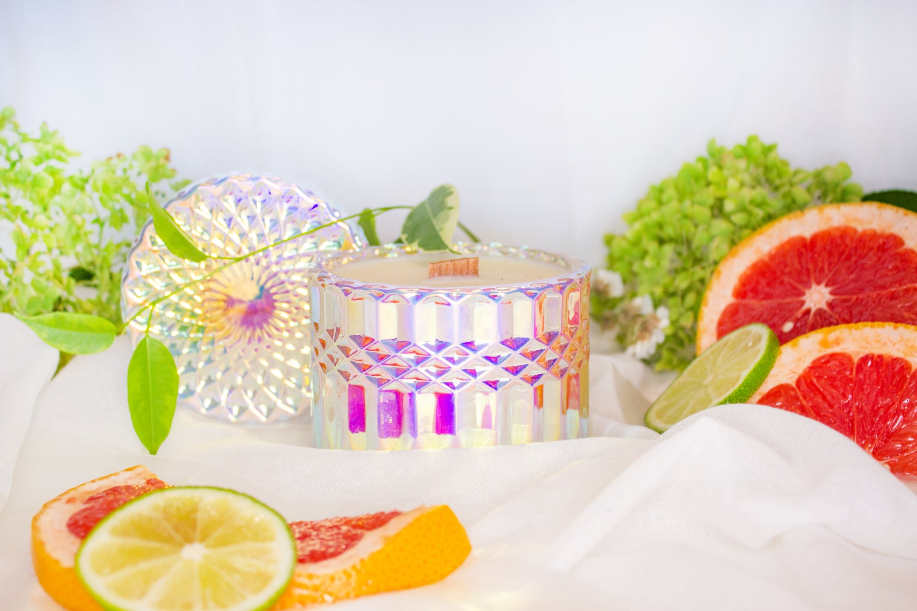 A candle in an iridescent cut glass jar is arranged among grapefruit slices and flowers