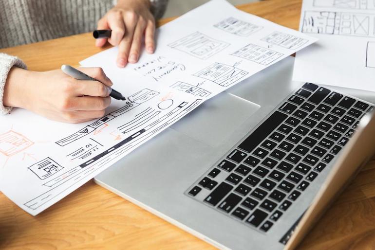 App store lessons: Photo of a web designer's hands drawing wireframes with a permanent marker over two large pieces of paper. A Macbook computer sits on the desk in front of the designer.