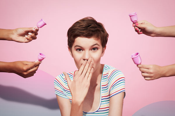 Foto de artigo sobre empreendedorismo feminino mostra uma mulher ao centro, com uma mão cobrindo a boca em uma expressão de choque. Ao redor da mulher, há quatro mãos diferentes, cada uma delas segurando um copo menstrual.