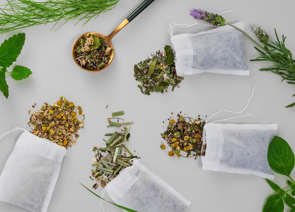 佛to para ilustrar artigo sobre vender produtos para mães mostra quatro sachês de chás sobre uma mesa, junto com uma concha que contém folhas de chá. Ao redor dos sachês, há folhas verdes de decoração.