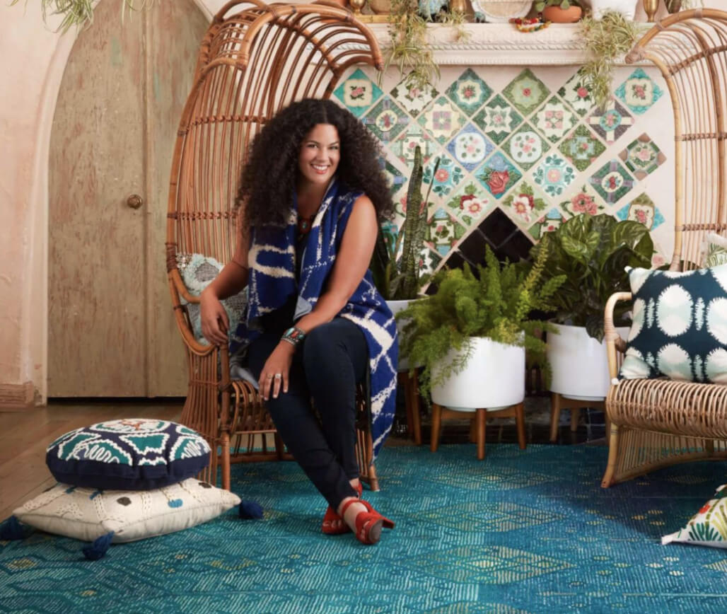 Founder of The Jungalow, Justina Blakeney, sits in a tall wooden chair in a room nicely decorated with many colourful pillows and an assortment of decorative potted plants.